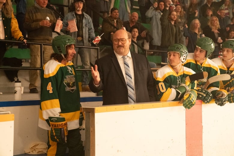 A still from a movie shows hockey players and a man in a suit on a bench in a hockey rink.