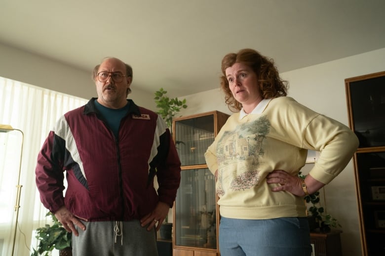 A still from a film shows an older man and a woman stand with their hands on their hips in a living room.