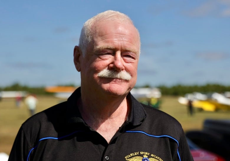 A man with white hair and a white mustache wears a black polo shirt. 