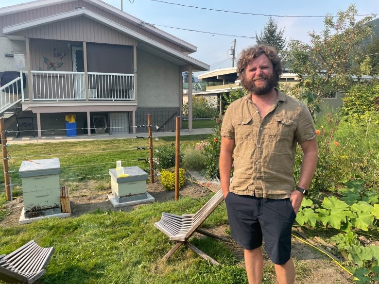 A bearded man in his backyard smiles for the photo. Patio furnitures, beehives, garden and a house can be seen in the background. 