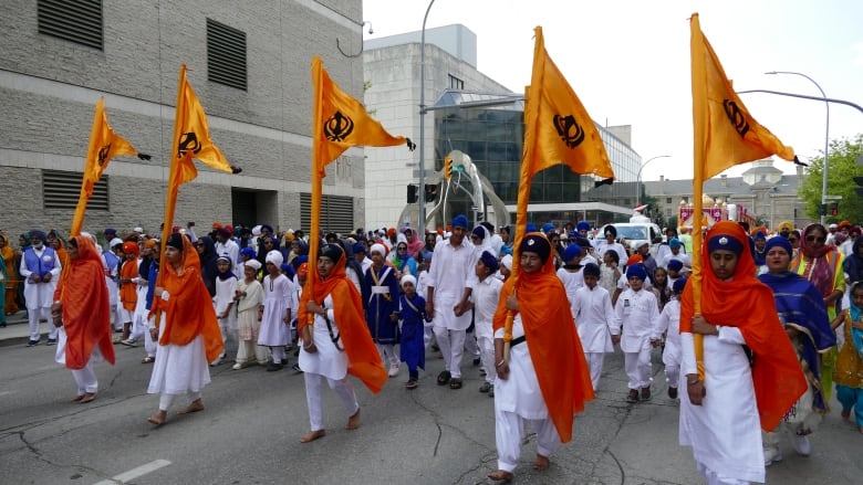 Hundreds of people walking on a street, led by people in orange and white bearing the Sikh flag