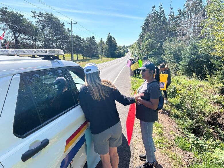 Two people holding protest signs speak to an officer in an RCMP vehicle.