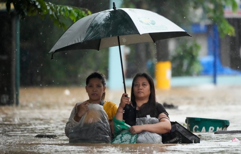 Two women proceed down a flooded street in chest-high water, clutching plastic bags. One holds an umbrella over their heads. 