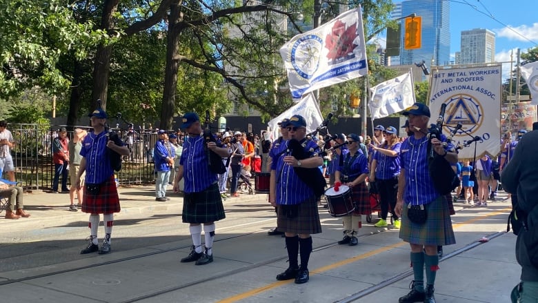 At a parade in downtown Toronto during daylight, pipers line up on the street, with flags behind them