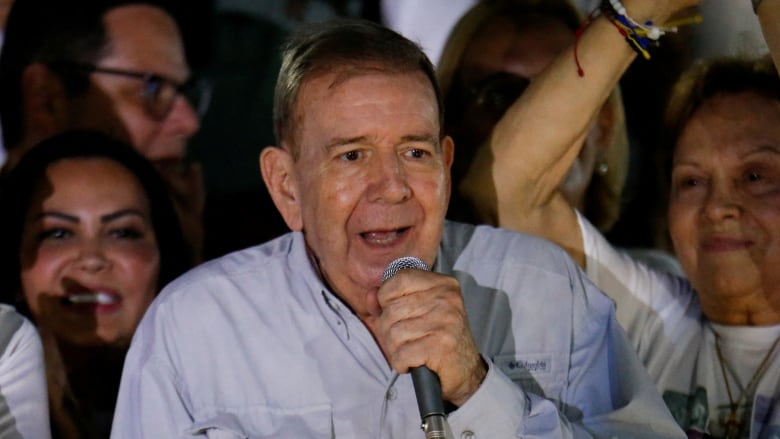 A person speaks while holding a microphone as people stand behind.