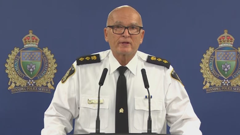 A bald man in a white shirt stands at a podium with microphones. 