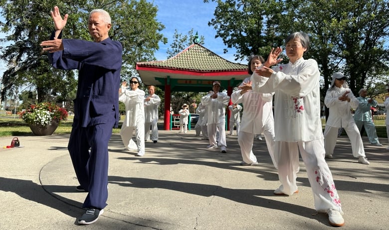 A group of seniors dressed in loose-fitting uniforms practice tai chi in a park with a Chinese pergola behind.