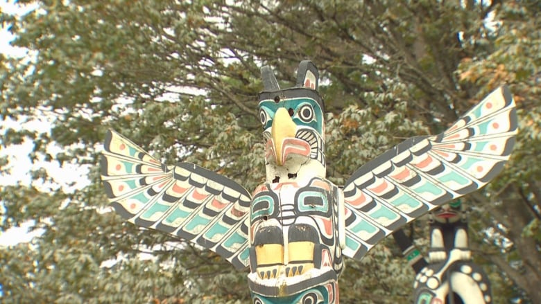 A thunderbird is seen atop a totem pole, with trees in the background.