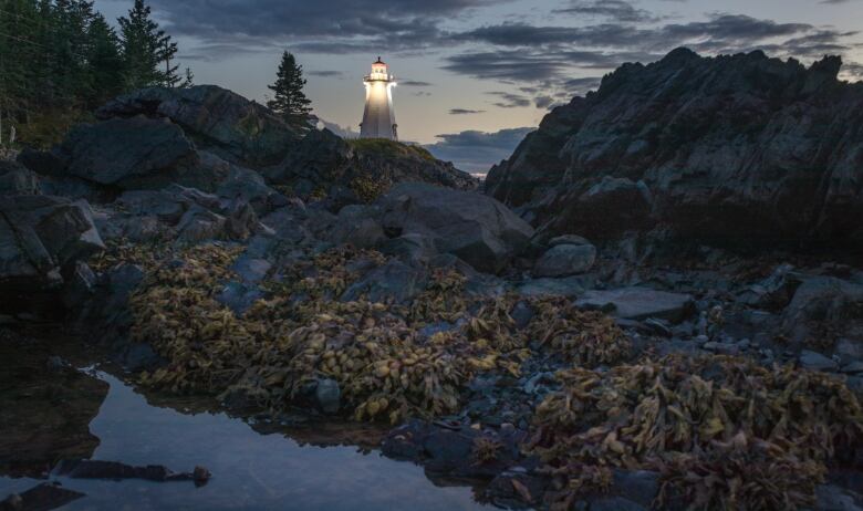 A lighthouse shines over a rocky island as the sky darkens.