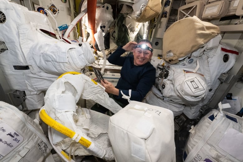 A smiling man with a blue shirt and a headlamp smiles at the camera with white cargo packs and spacesuits.