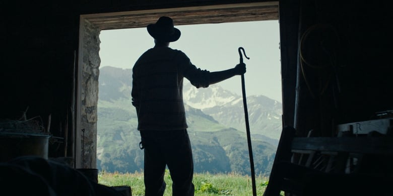 The silhouette of a man in a cowboy hat holding a staff stands in the door frame looking out at mountains.