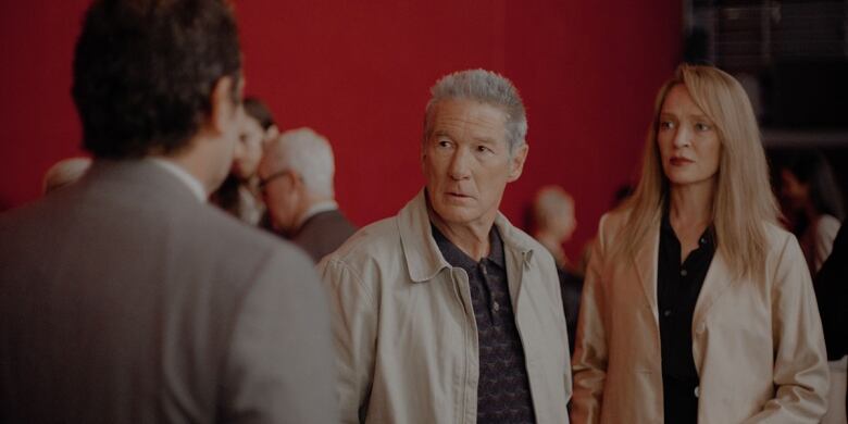 Richard Gere and Uma Thurman dressed in beige jackets stand in a room in front of a large red curtain.