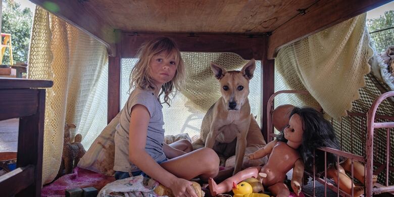 a young girl, a dog and a doll sit in a fort made of a table and blankets.