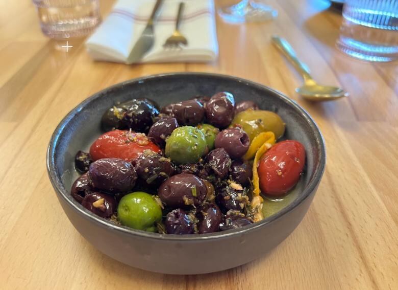 Dark brown, green and red olives sit in a dark bowl on a wood table.