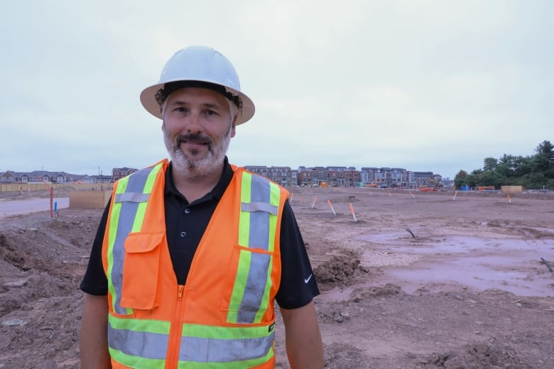 Man in hard hat and safety vest stands in front of a muddy plain with orange tipped pipes sticking out of it and houses in the distant background.