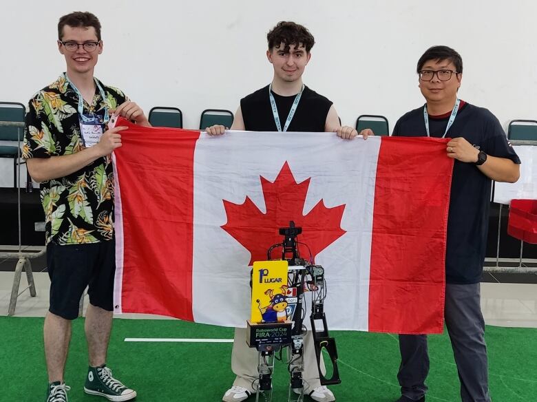 Three people holding a Canadian flag with a robot that looks like a small person in front of it.