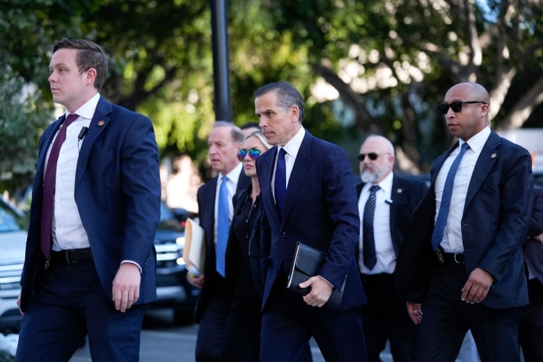 A man with dark hair in a blue suit walks into a courthouse surrounded by his wife, a blonde woman wearing sunglasses, and security agents in black suits.