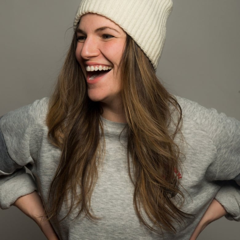 Woman with long brown hair, wearing a cream-coloured toque, puts her hands on her hips and laughs. 