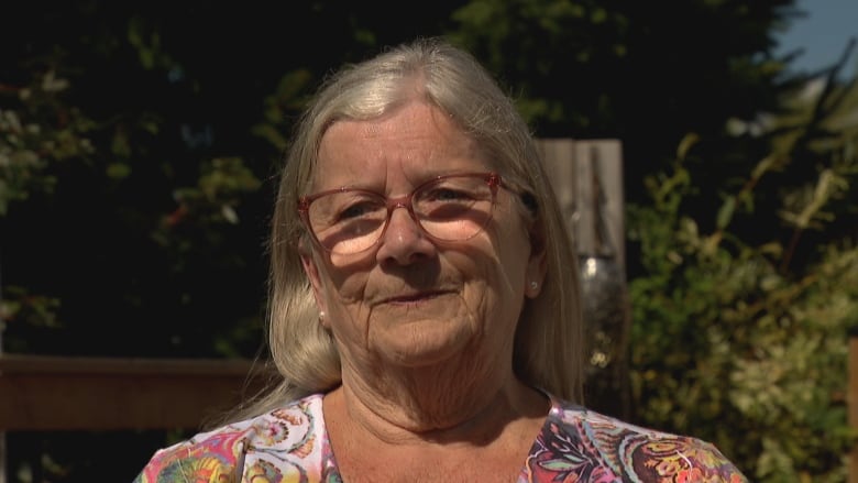 A woman with long white hair, parted in right of centre, wearing glasses and a pastel muli-coloured print shirt, looks towards the camera in full sun with pursed lips in a head and shoulders image.