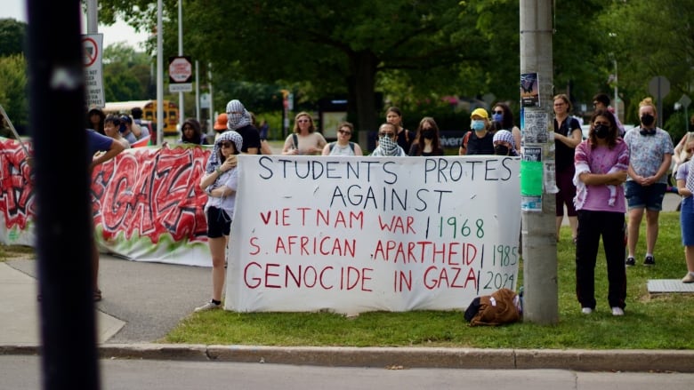 People stand in line on a street corner. Two hold a banner likening protests against Israel's actions in Gaza to protests agains the Vietnam War and South African Apartheid. 