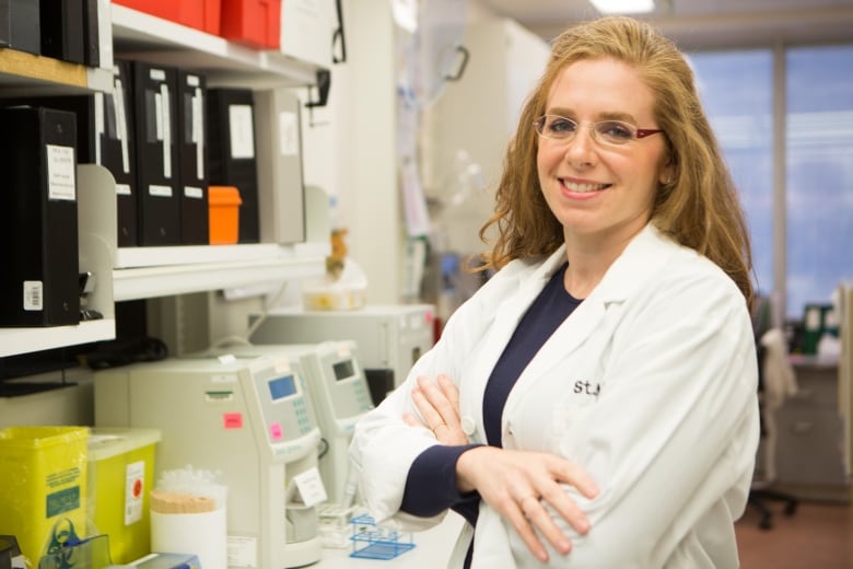 A woman in a lab coat stands with her arms crossed.