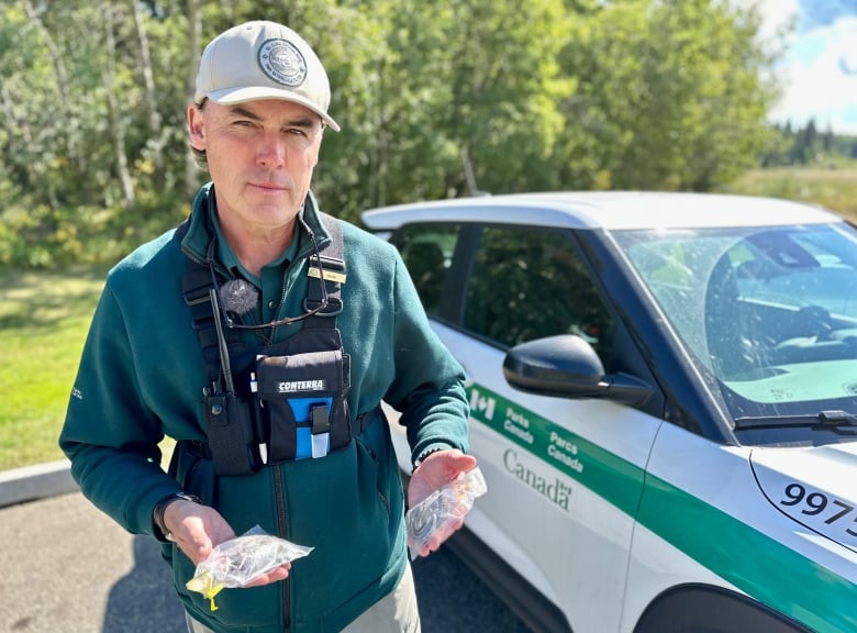 A man in a ball cap holds two clear plastic bags with two dead garter snakes, next to his  Parks Canada truck. 