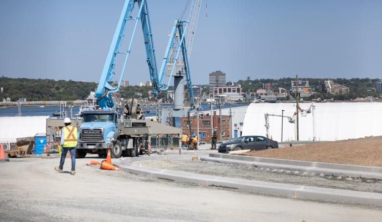 The edge of a mound of dirt surrounded by a gravel roadway and curbs is seen with a man in a high-vis vest walking away from the camera