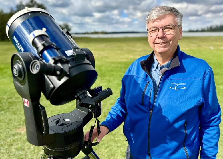 A smiling man in a blue jacket stands next to a telescope with some grass and a lake in the background.