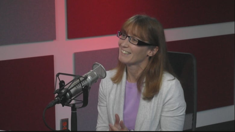 A woman with thick, square glasses sits in a radio studio in front of a microphone.