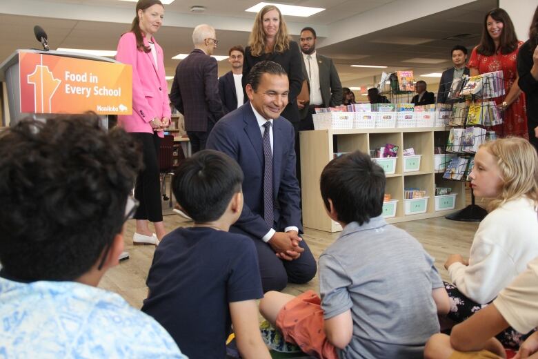 A man in a dark blue suit and tie kneels on the floor in front of a number of kids sitting on a rug.