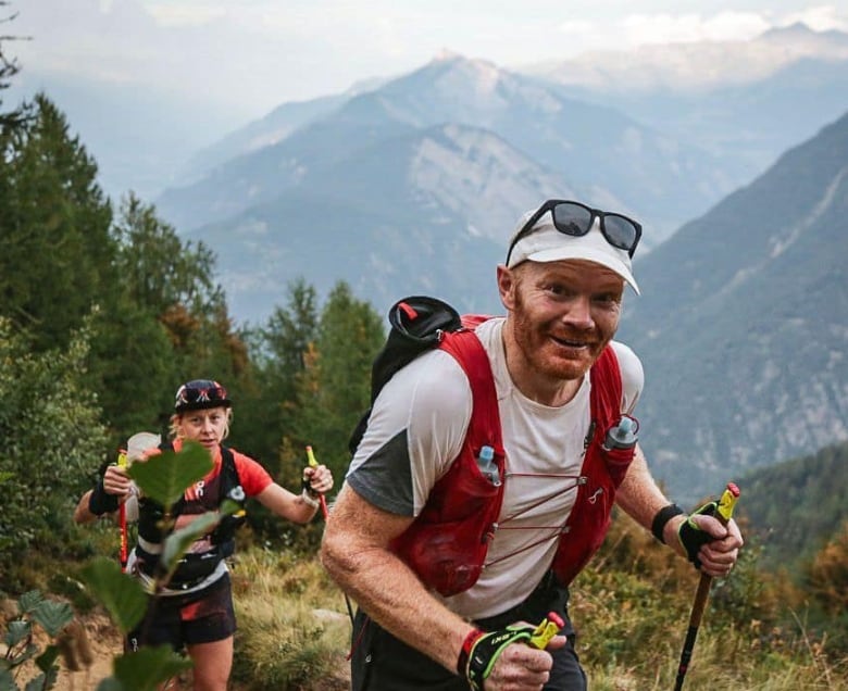 A man with red hair and a red beard is seen hiking up a mountain in Europe. He is smiling at the camera, and a woman is seen hiking behind him.