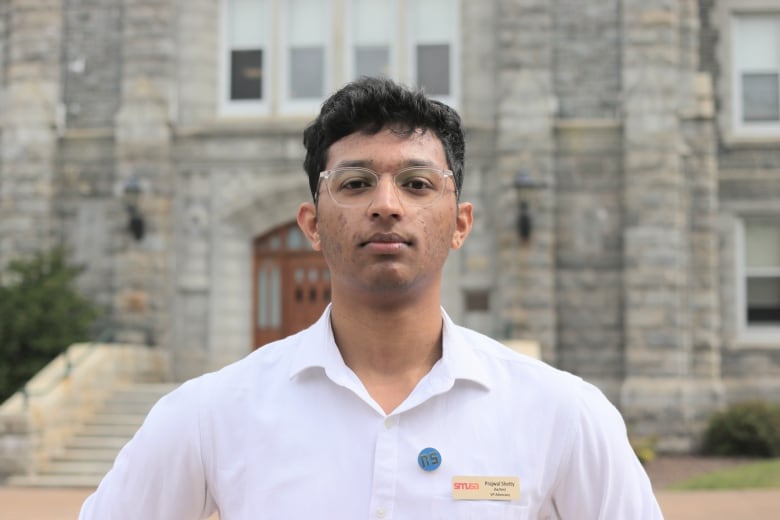 A person with short hair wearing glasses and a collared shirt stands in front of a stone school building with steps. 