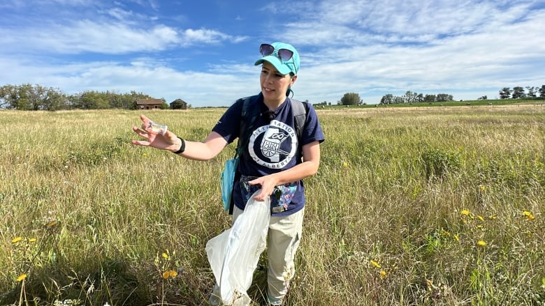 Woman wearing a navy blue shirt, tan pants, and a turquoise hat standing in a field, holding a vial with a bumblebee in it. 