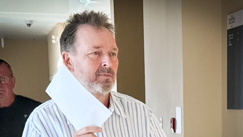 A man with greying hair, beard and moustache wearing a white shirt with blue stripes holds up a piece of paper to try to block his face.