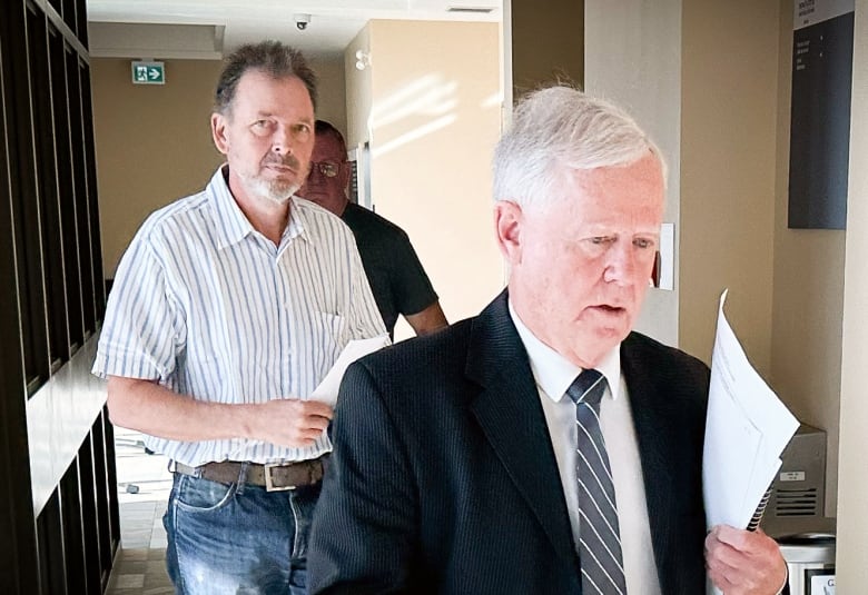 A man with greying hair, beard and moustache wearing a white shirt with blue stripes walks behind a white-haired man in a blue suit.