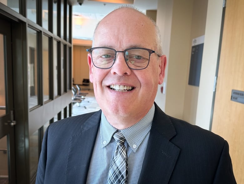 A balding man with glasses wearing a dark blue suit and light blue shirt and tie smiles as he stands in a hallway.