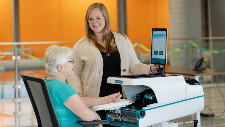 An older woman sitting next to a medical device as a smiling younger woman looks down.