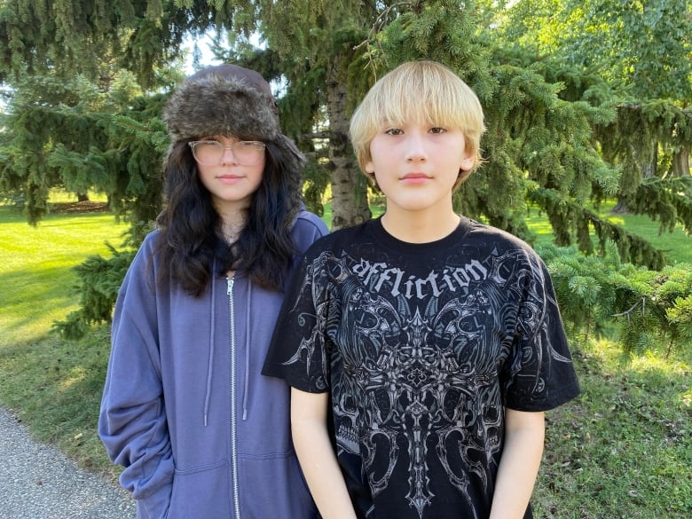 Two teens stand looking serious in front of a tree.