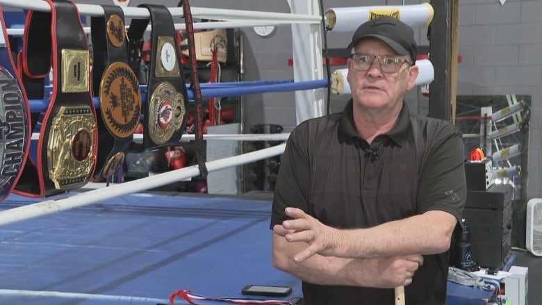 A man wearing a black shirt and a black cap stands next to a boxing ring with various championship belts draped along the ropes.