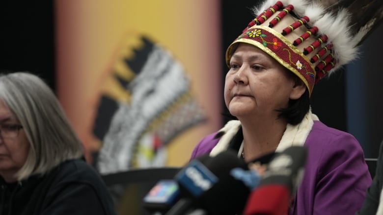 A woman wearing an Indigenous feather headdress sits and looks forward, with microphones in front of her.