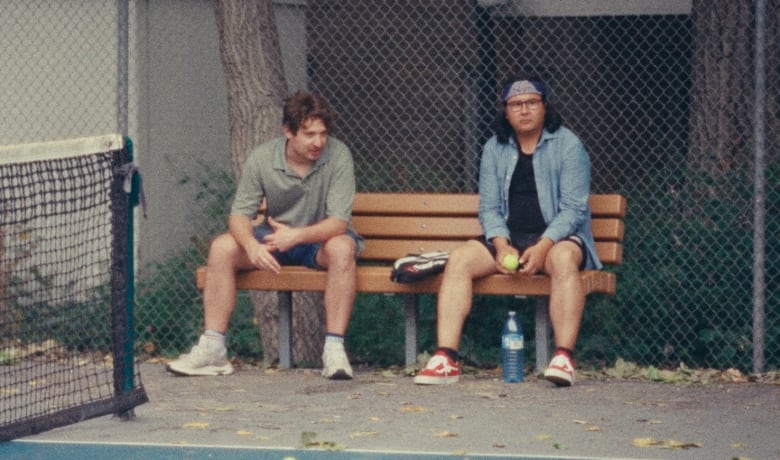 A still from a movie shows two men sitting on a bench next to a tennis court.