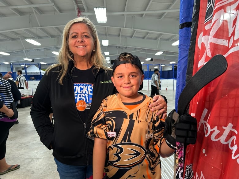 A woman with long blonde hair stands next to her son who's holding a hockey stick and wearing a hockey jersey.