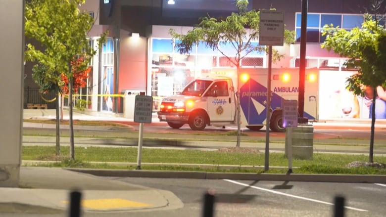 An ambulance with flashing lights is parked in a parking lot next to a business. Police tape surrounds the business. It is night time.