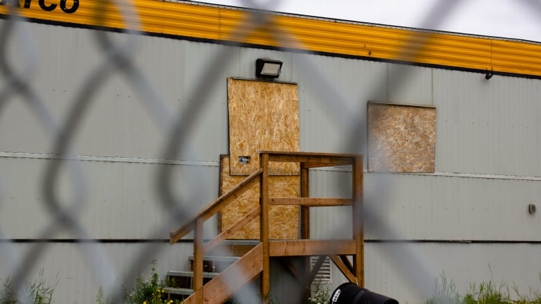 A grey trailer has its door and front window boarded up. It sits behind a chain link fence. A black garbage receptacle is tipped over in front of the front stairs.