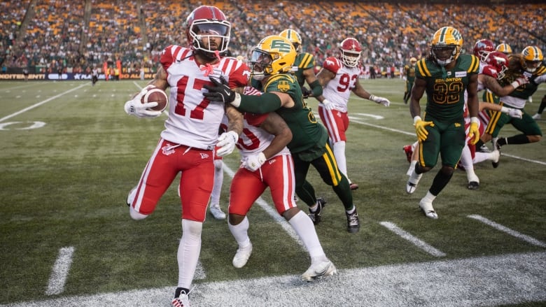 A football player in a red-and-white uniform carries a football while running onto the sideline, avoiding getting hit by nearby opponents in green-and-yellow uniforms.