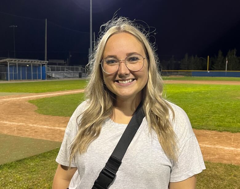 A woman smiles. She stands on a baseball field.