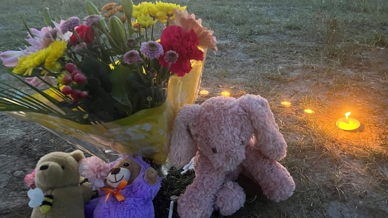 Three stuffed animals sit on grass in front of a colourful bouquet. A handful of tealight candles are lit nearby.