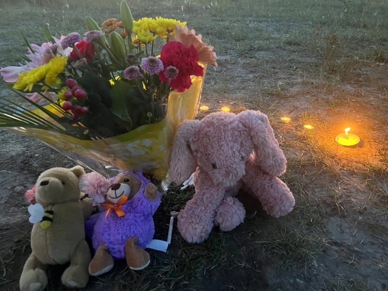 Three stuffed animals sit on grass in front of a colourful bouquet. A handful of tealight candles are lit nearby.