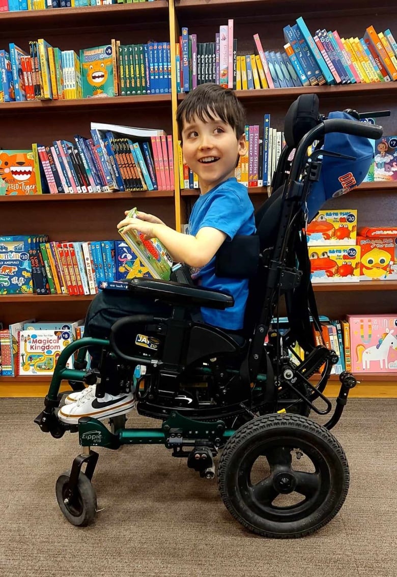 Image of a small boy in a wheelchair next to a bookshelf holding a book, smiling.