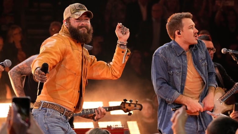 A bearded singer in an orange jacket and jeans dances onstage next to another male performer in a denim top and jeans.